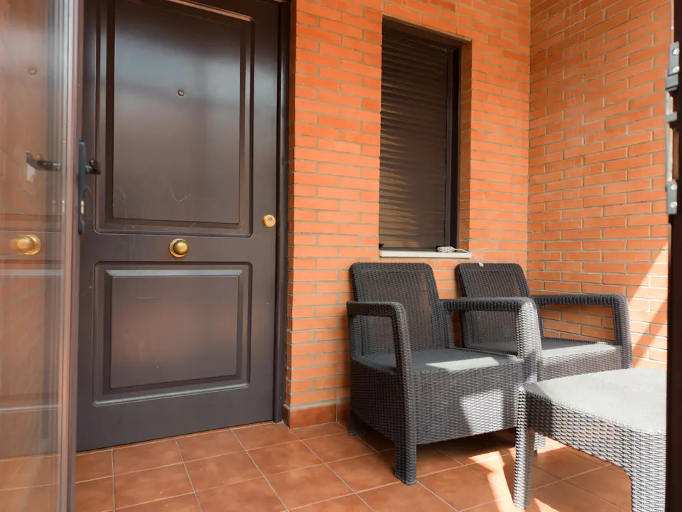 Outdoor patio with brick wall, dark door, and window with closed blinds. Two dark wicker chairs and a small table sit on the tiled floor.