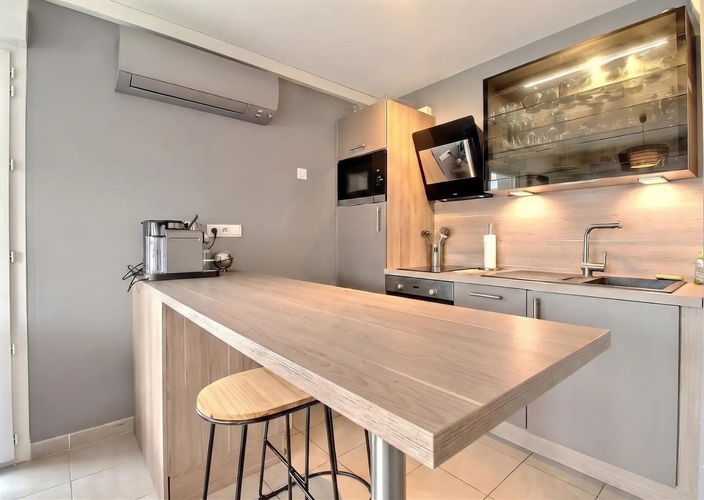 A modern kitchen with gray walls, light wood cabinets, and a breakfast bar with a stool. Appliances include a microwave, oven, and coffee maker.