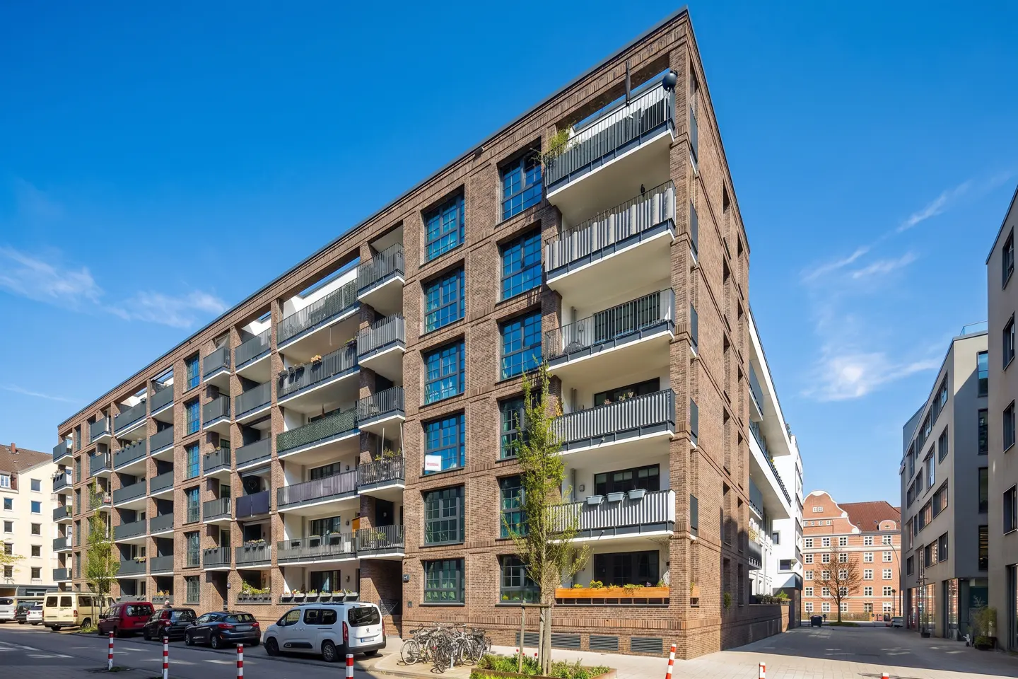 Exterior view of a modern brick apartment building with balconies under a clear blue sky. Cars and bikes are parked on the street.