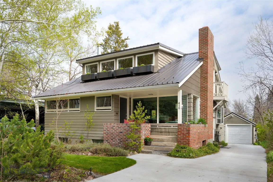 A two-story house with a metal roof, a brick chimney, and a small balcony. The house is surrounded by trees and greenery.