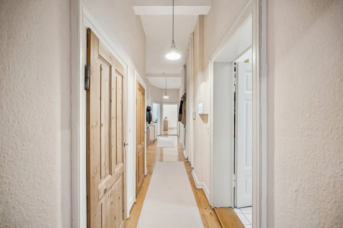 Hallway view with light beige walls, wooden floors, and doors. A white runner stretches down the center, leading to rooms illuminated by pendant lights.