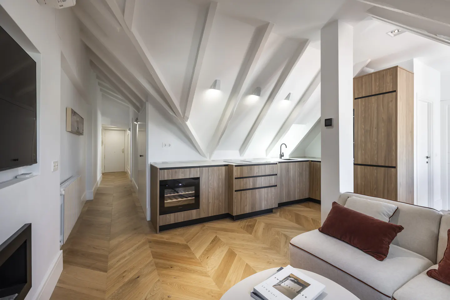 Bright, modern attic apartment with white walls, wood floors, and exposed beams. A small kitchen area and a cream-colored sofa are visible.