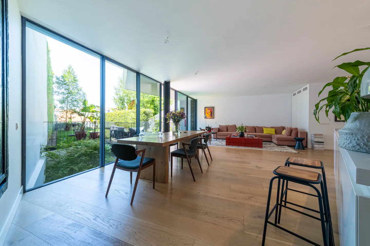 Modern open-plan living area with a wooden dining table, black chairs, and a beige sectional sofa overlooking a garden.