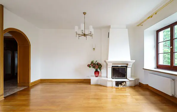 Bright, empty living room with wood floors, white walls, and a fireplace. A chandelier hangs from the ceiling. A large window lets in natural light.