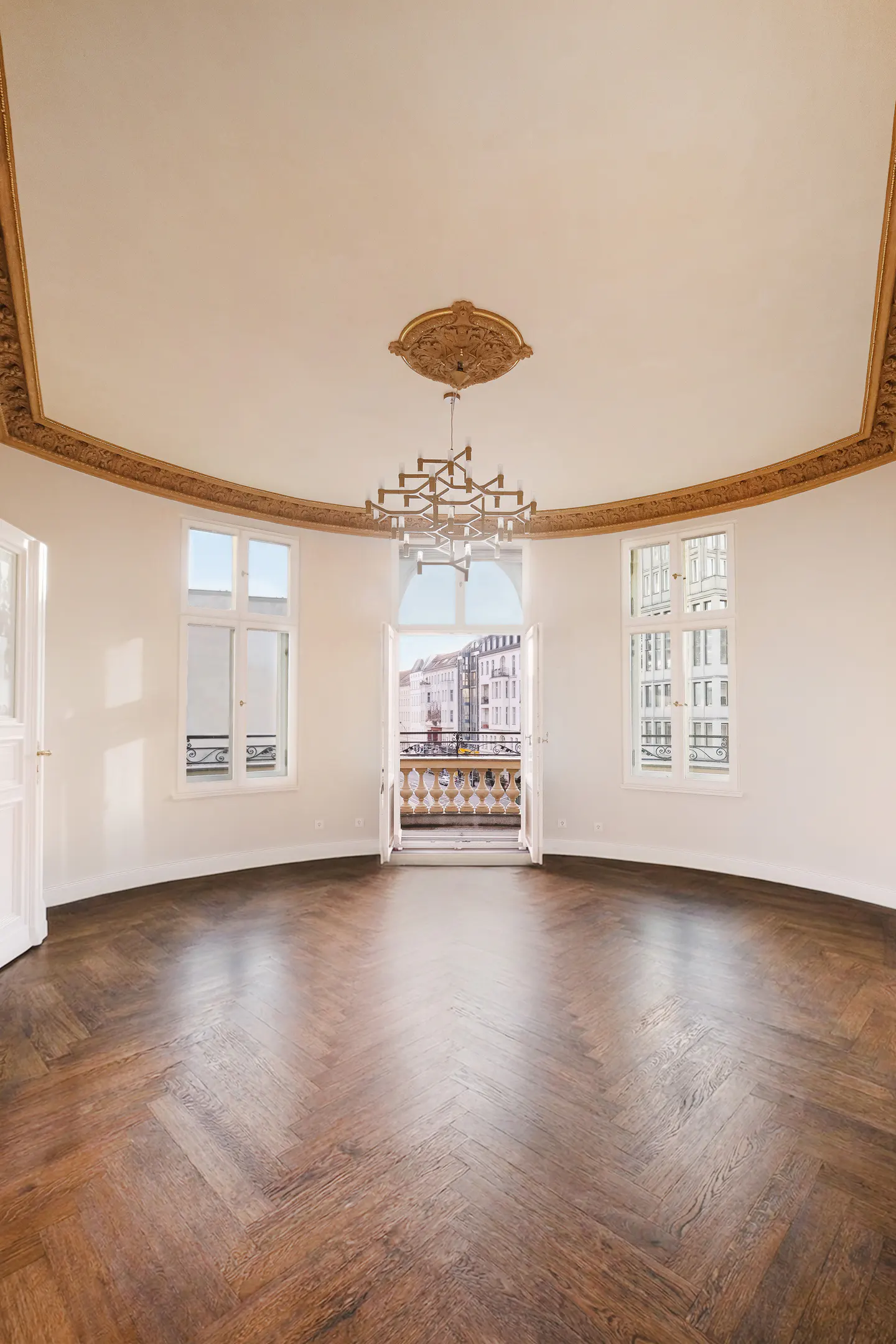 Bright, empty room with herringbone wood floors, white walls, and ornate gold trim. Balcony doors open to a city view. Chandelier hangs from the ceiling.