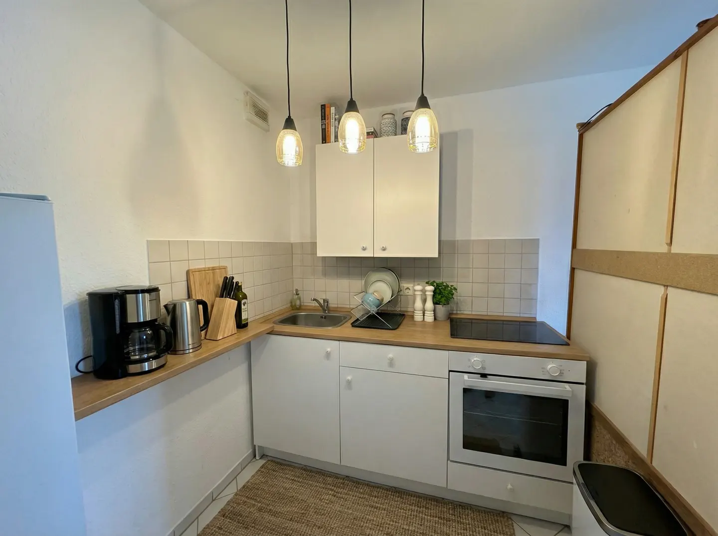 A bright, compact kitchen with white cabinets, wood countertops, and stainless steel appliances. Three pendant lights hang above.