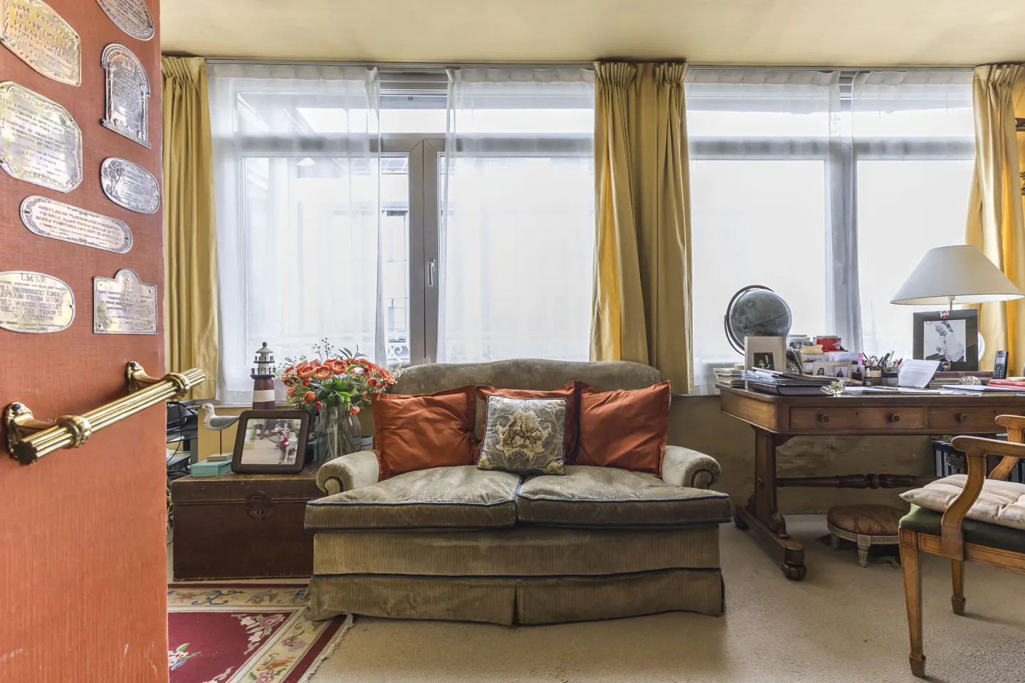 Cozy living room with a tan sofa, orange pillows, and a wooden desk by the window with yellow curtains. A red door is on the left.