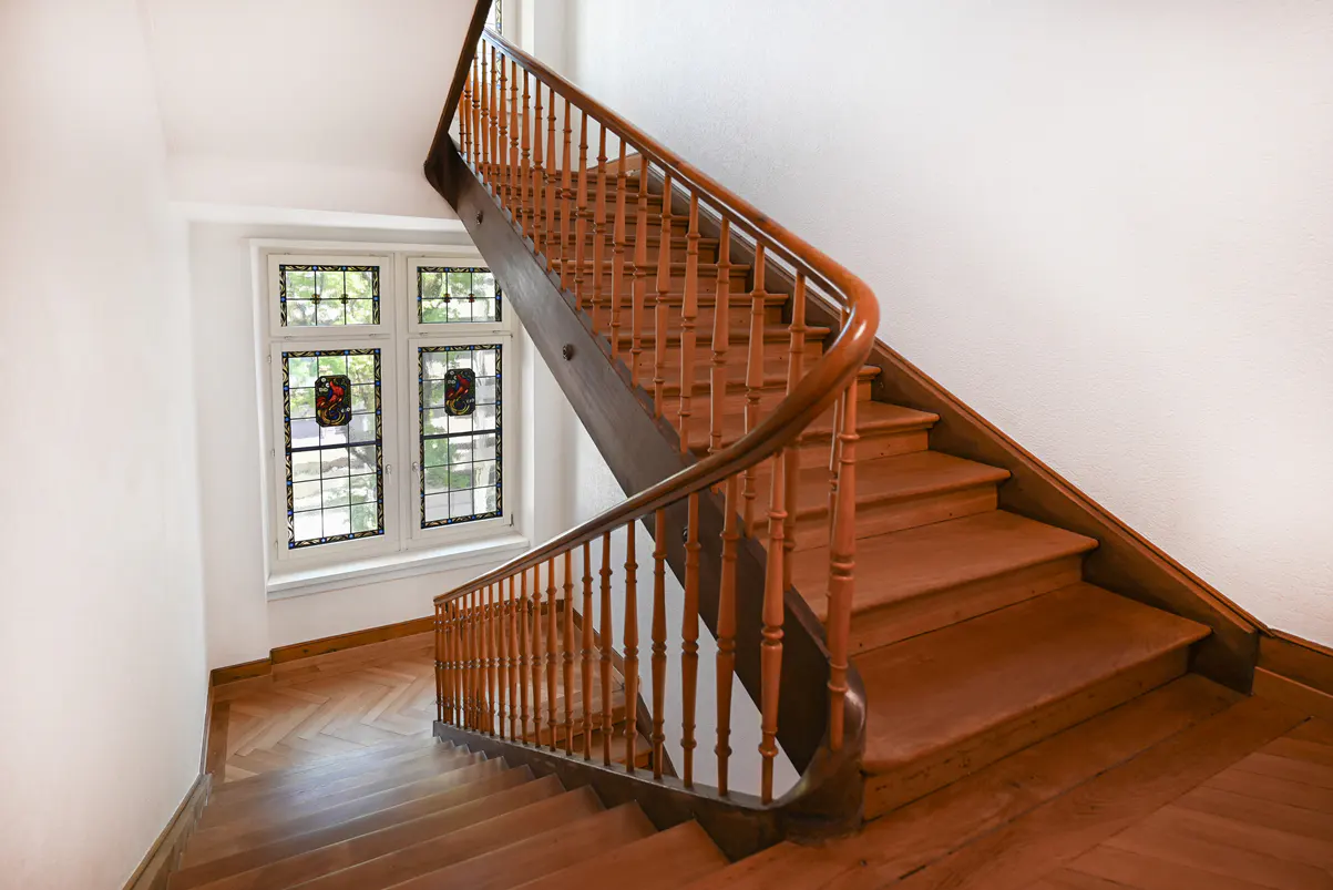 Wooden staircase with curved banister in a white-walled hallway. A window with stained glass is visible.