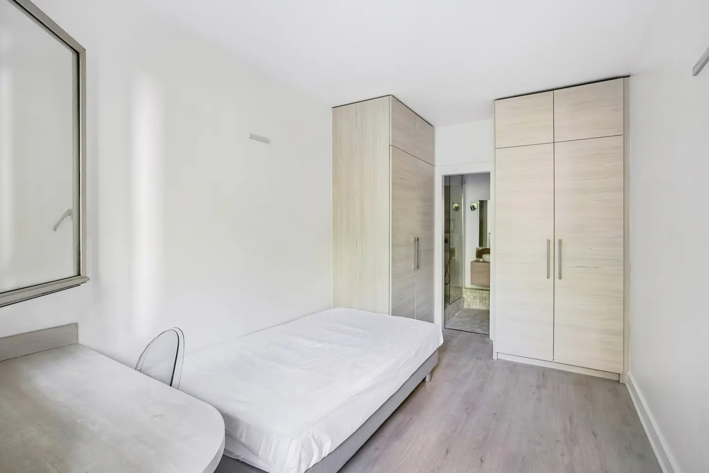 A bright bedroom with a single bed, desk, and two light wood wardrobes. A bathroom is visible through a doorway.