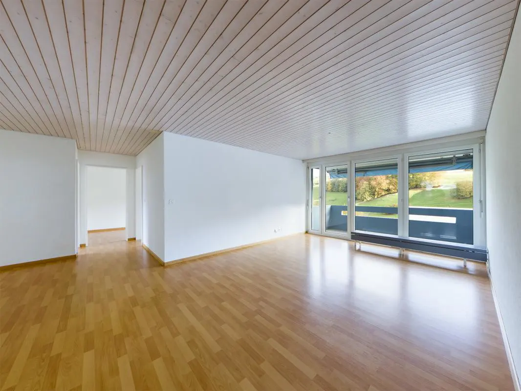 Empty room with light wood flooring, white walls, a slatted wood ceiling, and large windows overlooking a green landscape.