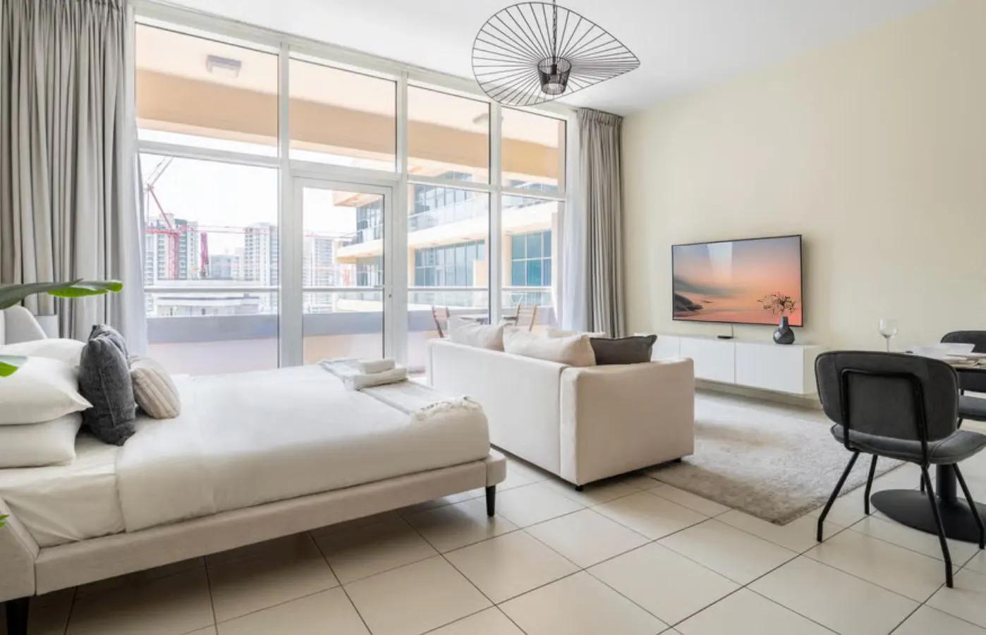 Bright studio apartment with a bed, sofa, TV, and dining table. Large windows offer a city view. Neutral color scheme with tile flooring.
