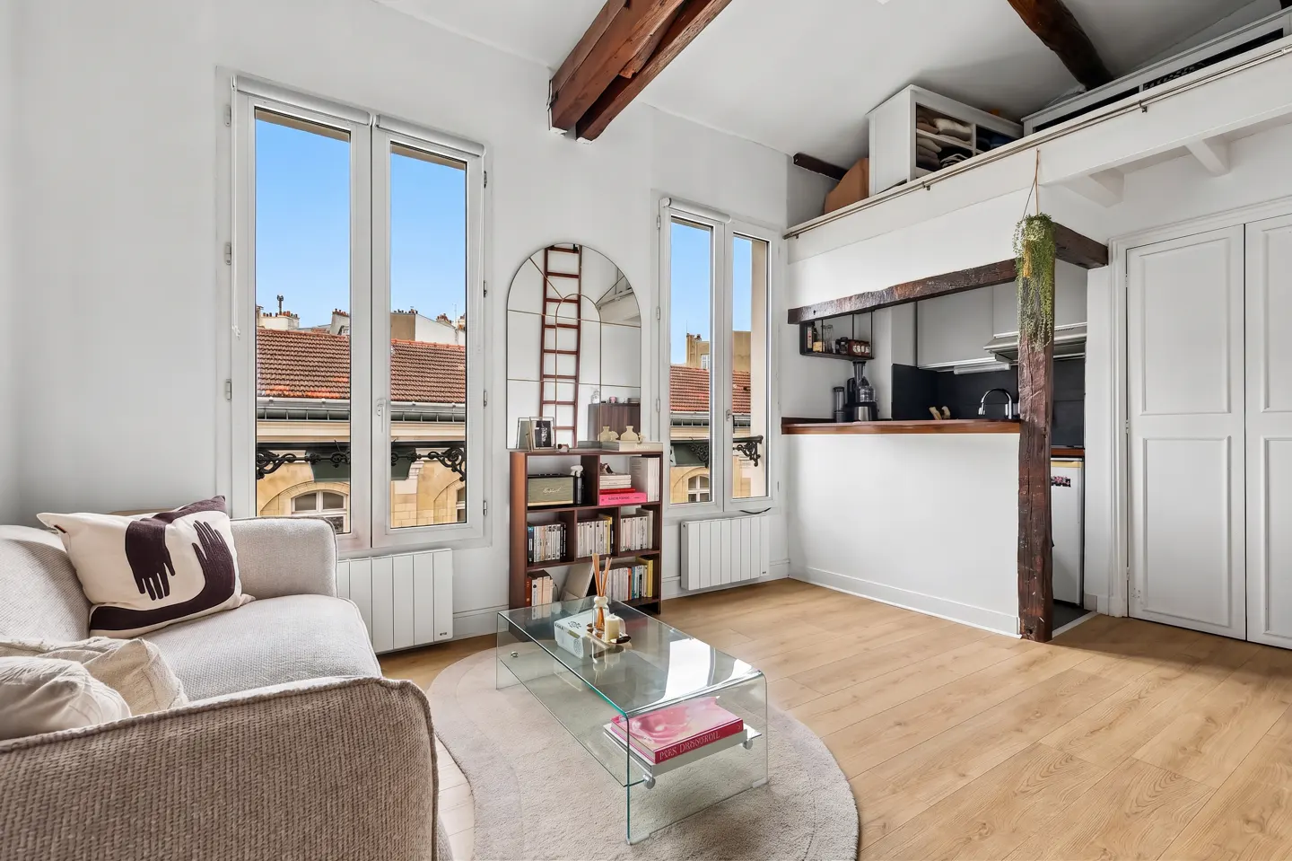 Bright apartment interior with wood floors, white walls, and exposed beams. A sofa, glass table, and kitchen area are visible. Windows show a cityscape.