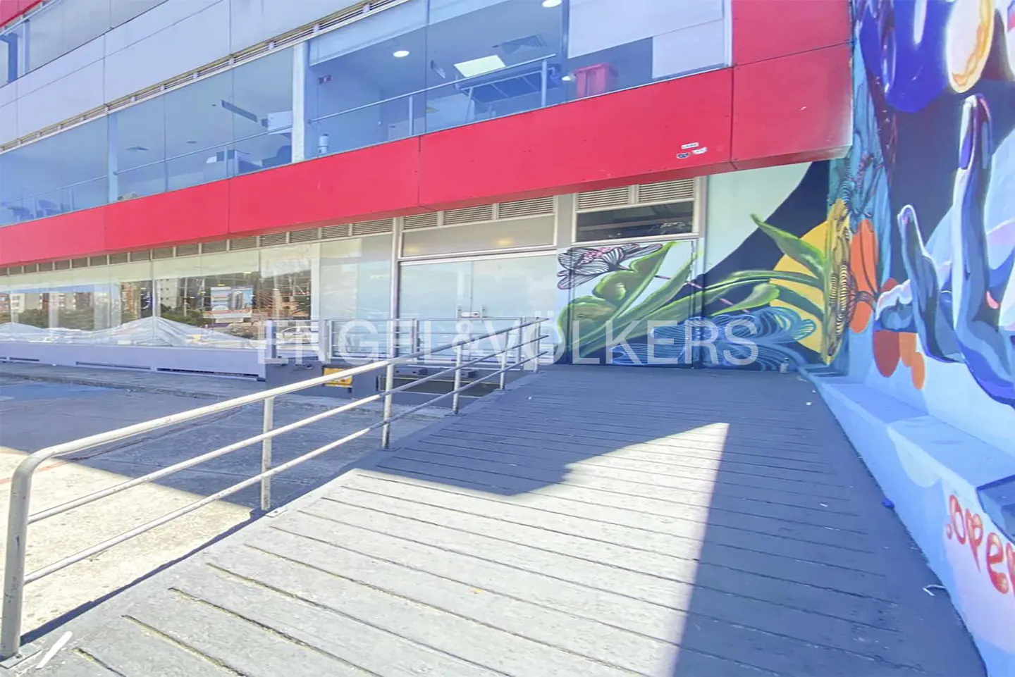Exterior view of a two-story building with a red accent and a ramp leading to the entrance. A colorful mural adorns the side of the building.