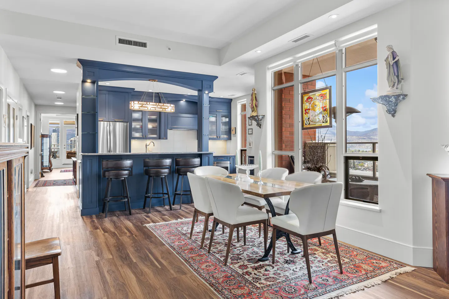 A bright, modern dining area with a blue kitchen island and bar stools. A table with white chairs sits on a patterned rug near a large window with a view.