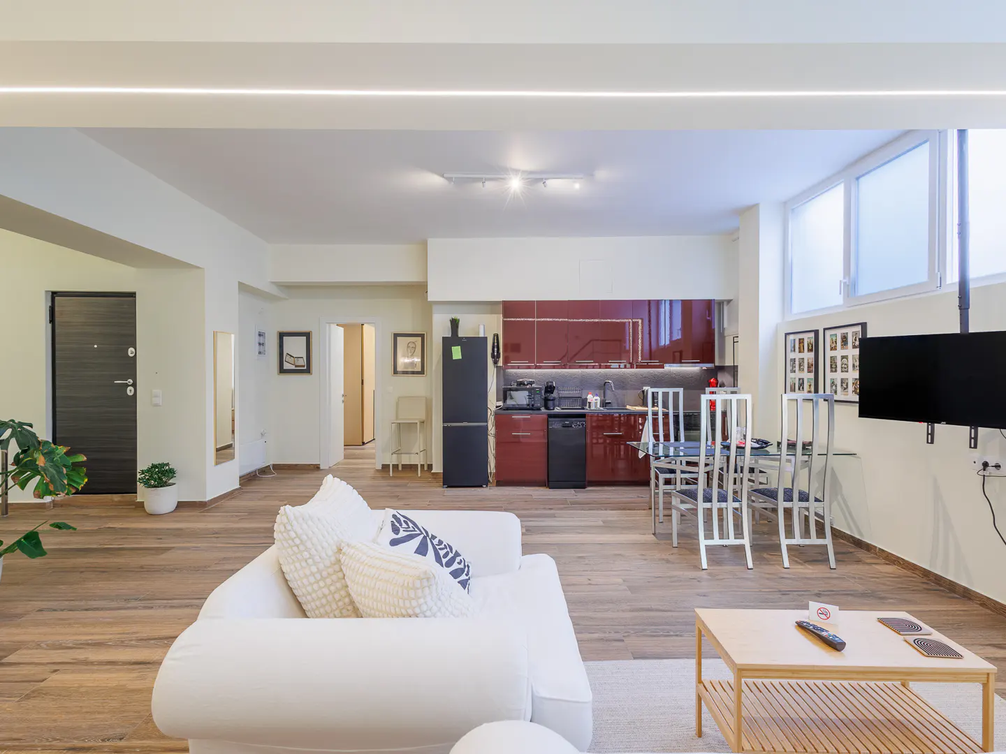 Bright, open-concept apartment with wood floors, white sofa, and red kitchen cabinets. A black refrigerator and TV are also visible.