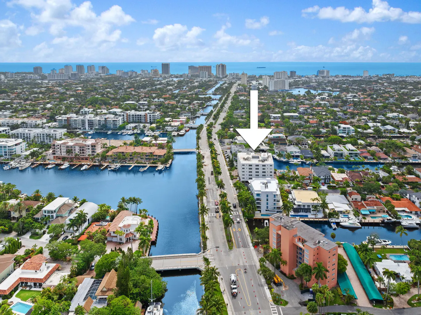 Aerial view of a white apartment building with an arrow pointing to it, located on a street lined with palm trees and canals.