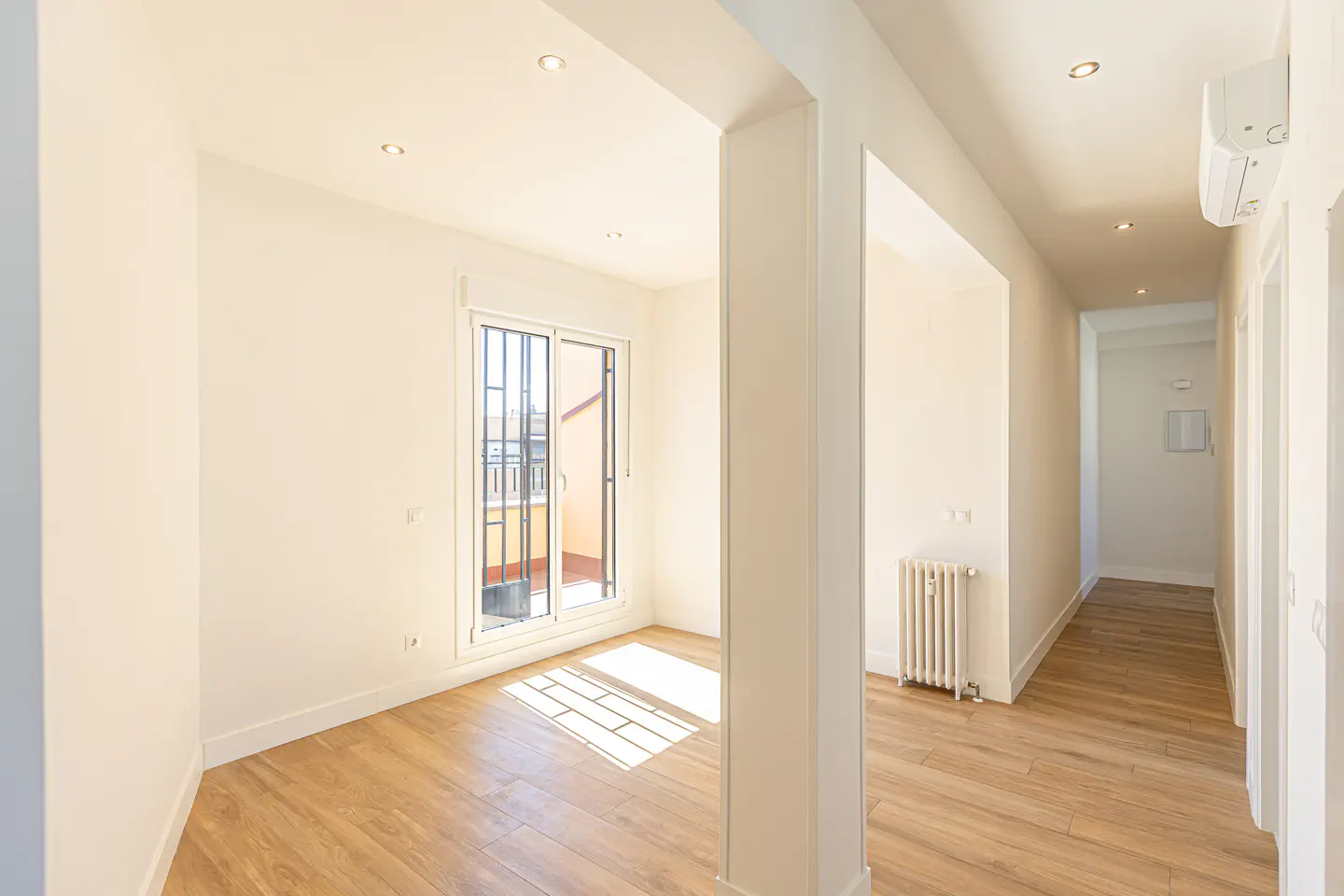 Bright, empty room with wood floors, white walls, and recessed lighting. A sliding glass door leads to a balcony. A white radiator is visible in the hallway.