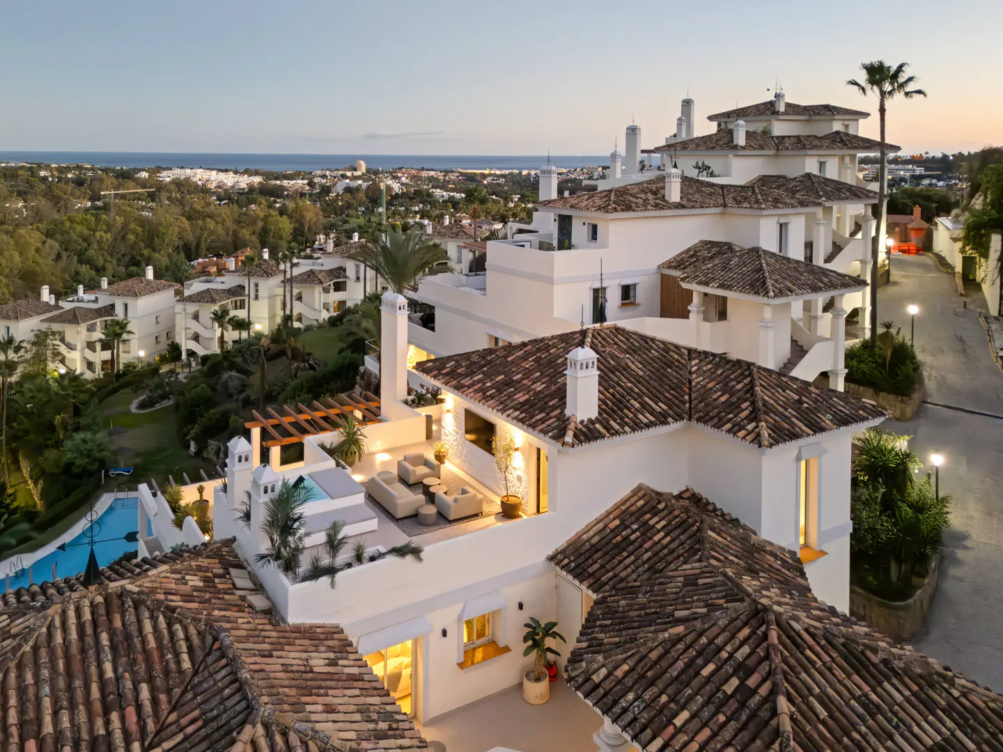 Aerial view of a multi-story white building with terracotta tile roofs, a rooftop lounge area, and a distant ocean view.