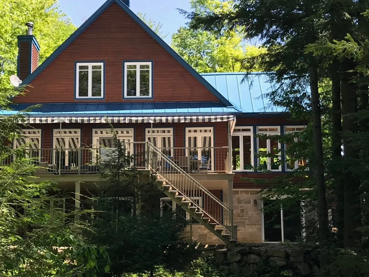 Two-story house with brown siding, blue roof, and white trim, surrounded by green trees. A metal staircase leads to a second-story balcony.