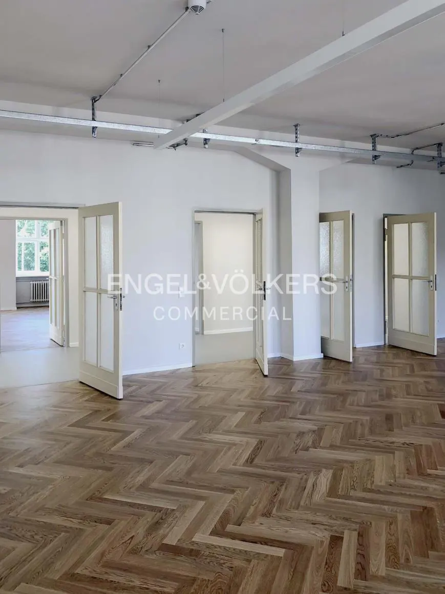 Empty commercial space with herringbone wood floors, white walls, and open doors. Engel & Völkers logo visible.