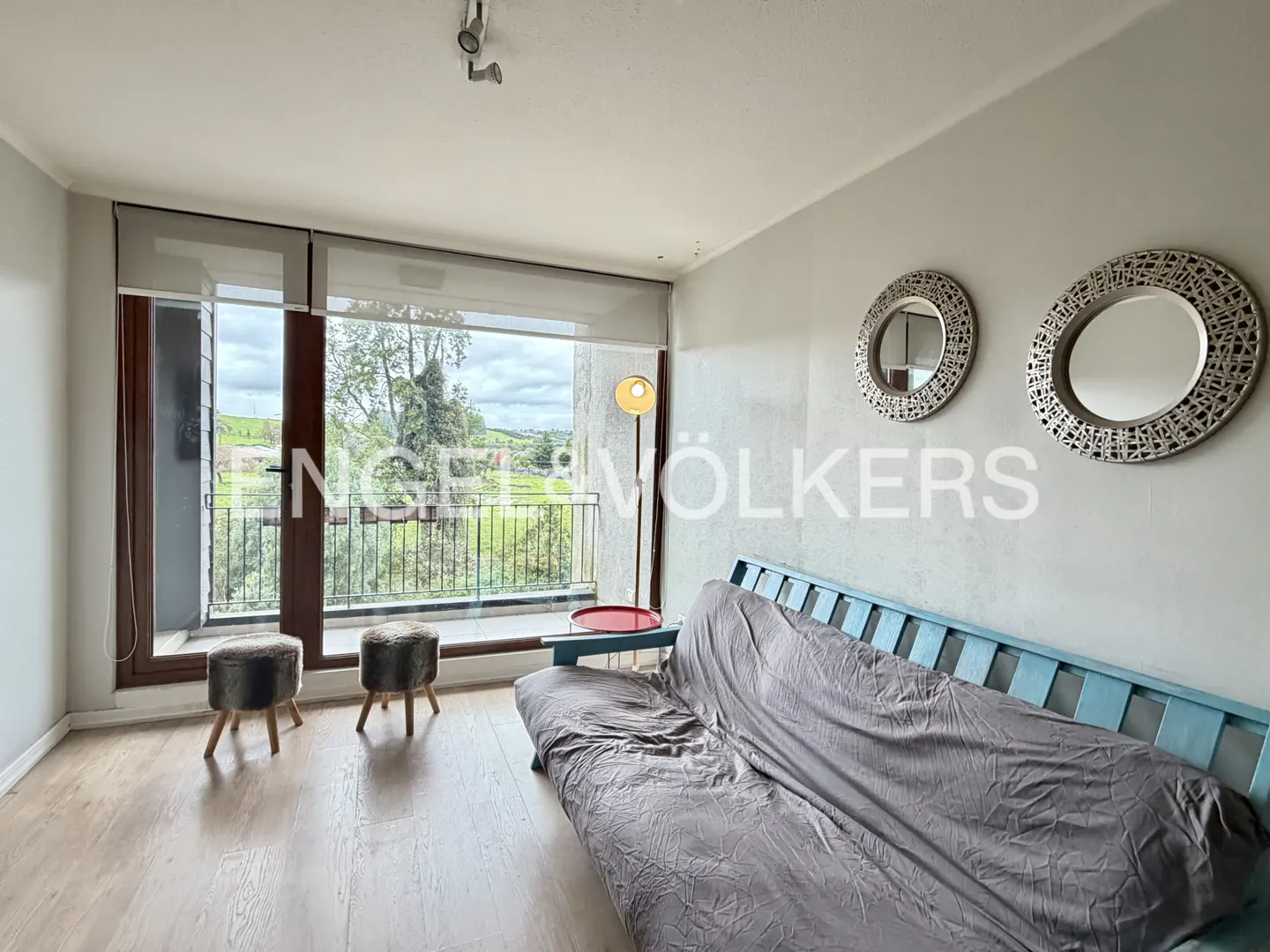 A bright living room with a blue sofa bed, two stools, and two round mirrors on the wall. A large window overlooks a green landscape.