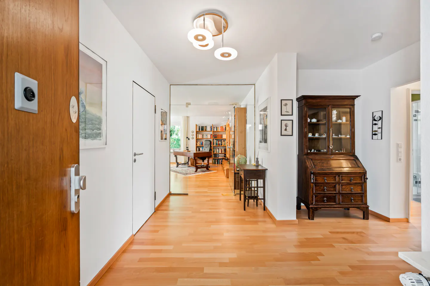 A bright hallway with light wood floors, white walls, and a wood door on the left. A desk and bookcase are visible through a mirror at the end of the hall.