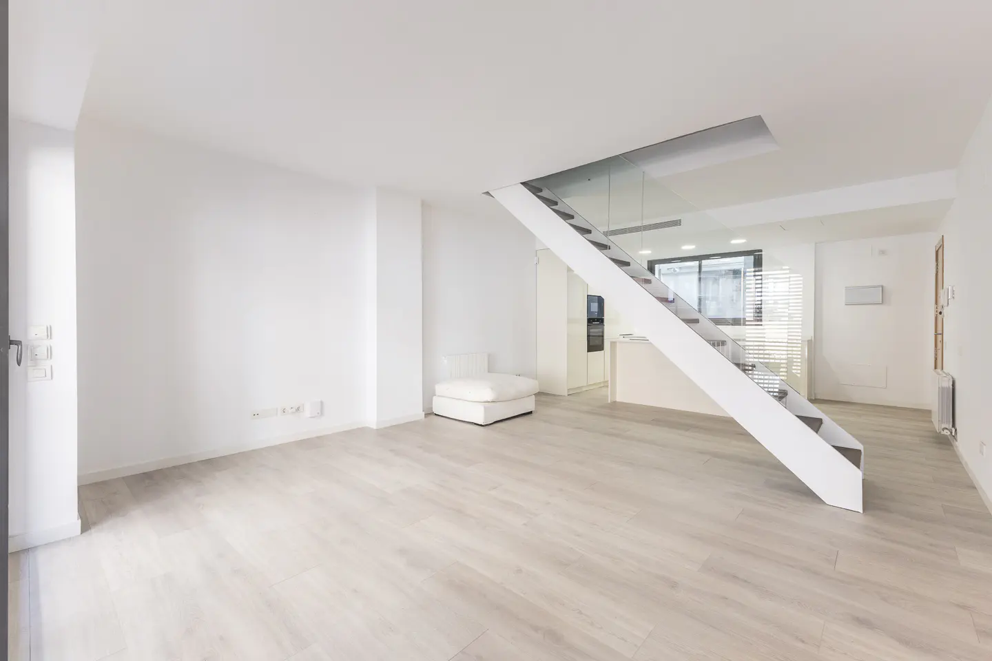 Bright, modern living room with white walls, light wood floors, and a white staircase with glass railing. A white ottoman sits in the corner.