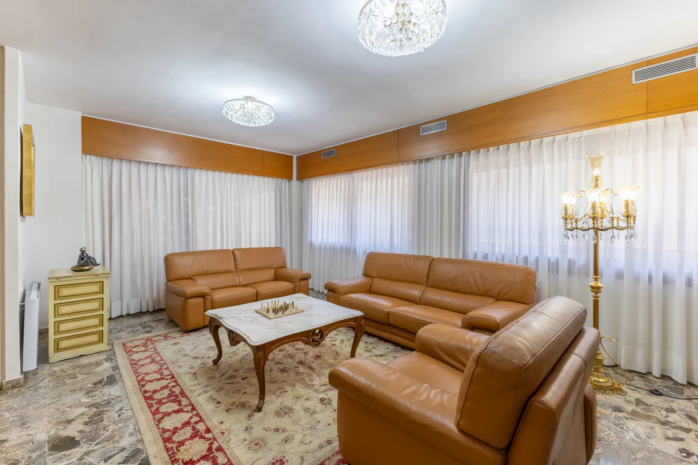 Living room with brown leather sofas, marble table with chess set, and crystal chandeliers. Cream rug with red pattern.