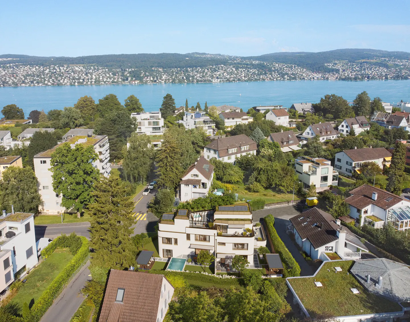 Aerial view of a modern white house with a pool, surrounded by trees and other houses, with a lake in the background.