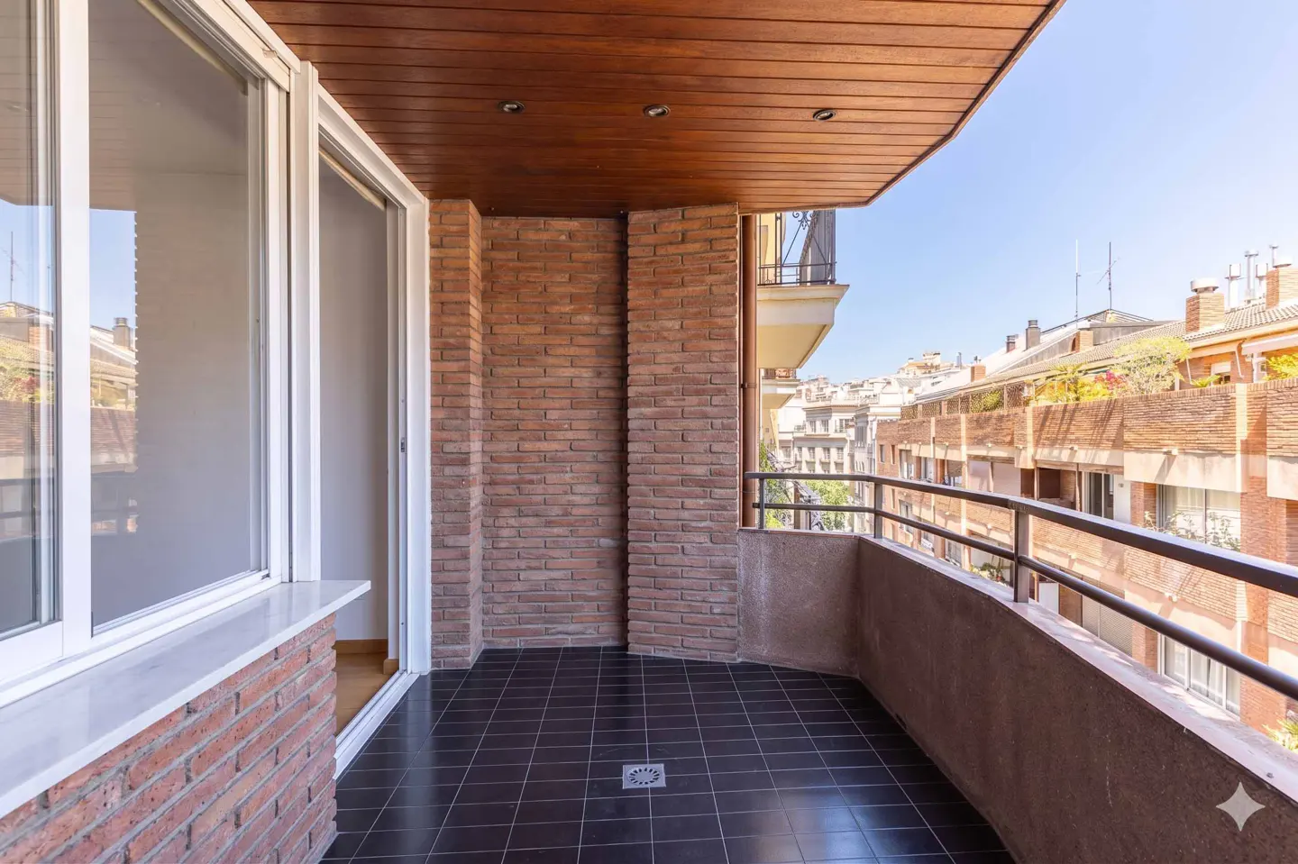 A balcony with brick walls, a dark tile floor, and a wooden ceiling overlooks a city street.