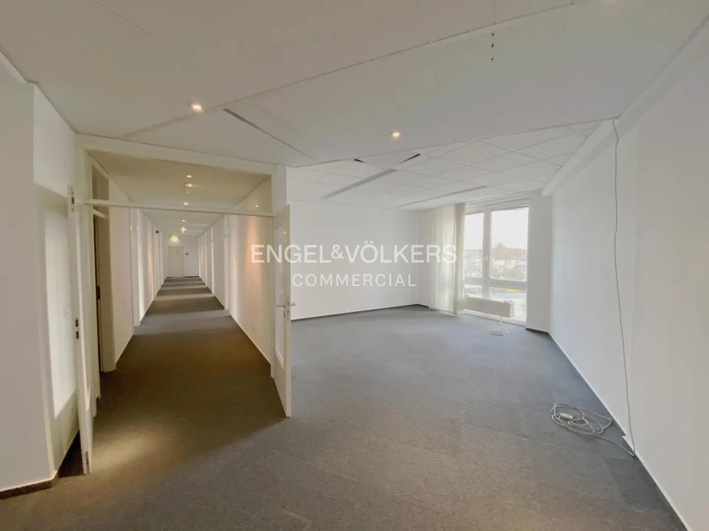 Bright office space with white walls, gray carpet, and a long hallway with doors. Engel & Völkers Commercial logo is visible.