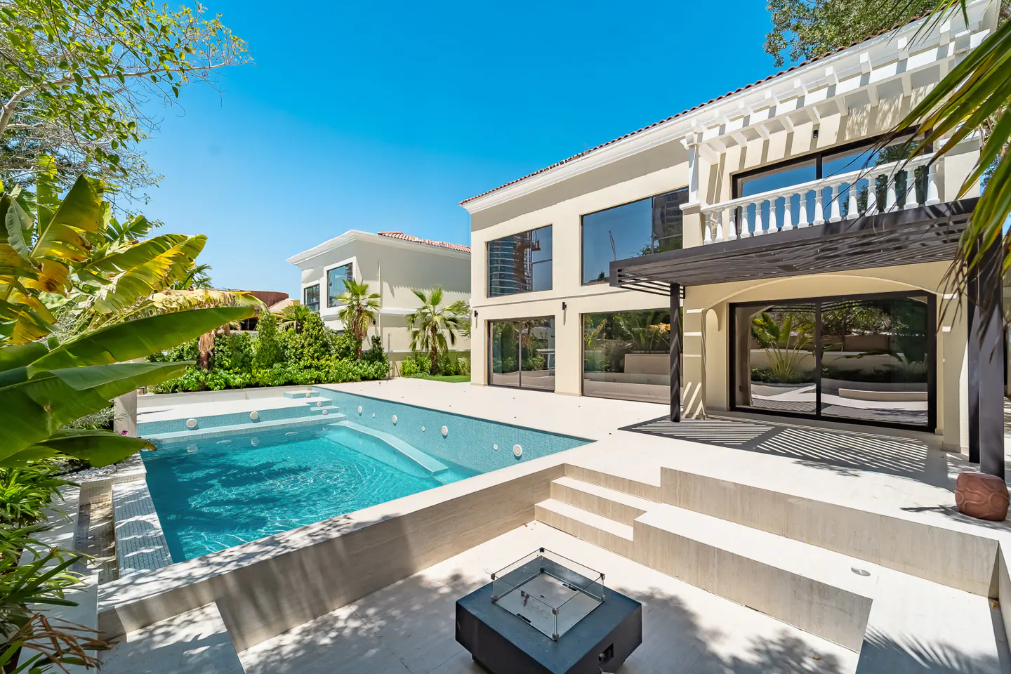 Backyard view of a two-story beige house with a pool, patio, and a black fire pit.