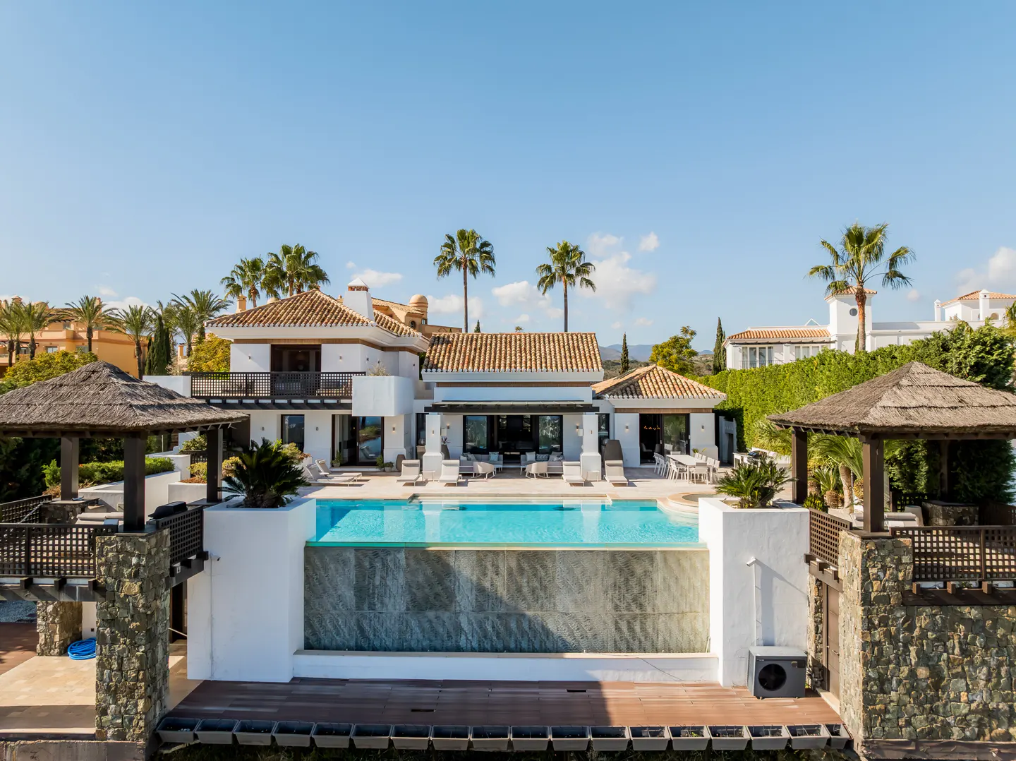 Luxury white villa with a tiered infinity pool, stone pillars, thatched gazebos, and palm trees under a clear blue sky.