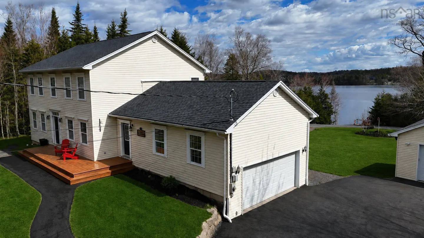 Two-story cream house with a black roof, a wooden deck with red chairs, and a view of a lake.