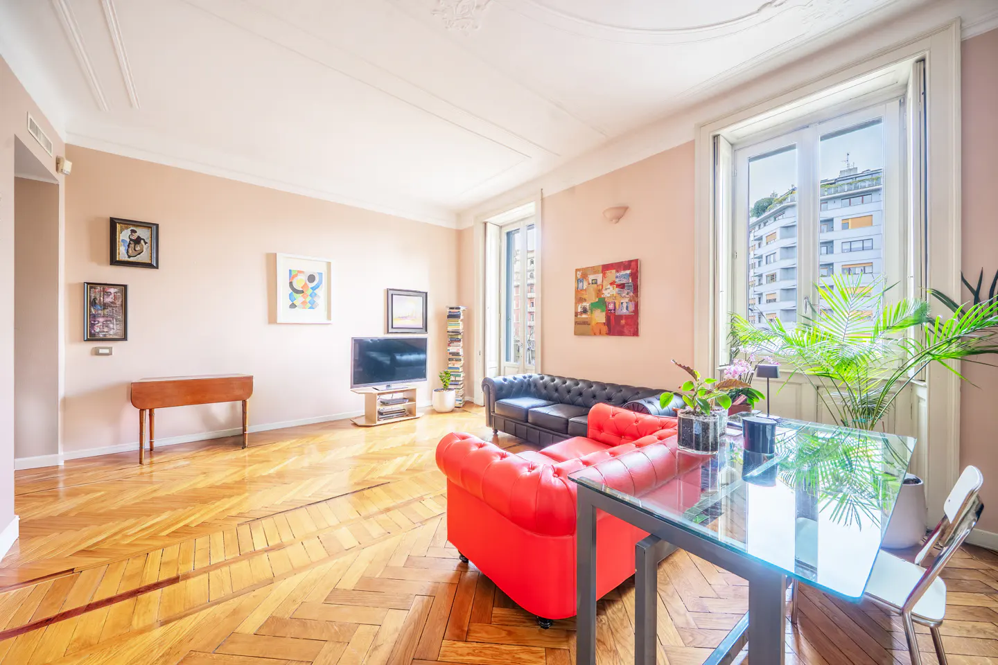 Bright living room with herringbone wood floors, pink walls, and large windows. Red and black leather sofas, glass table, and artwork decorate the space.