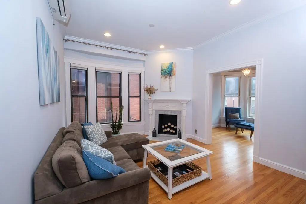 Living room with brown sectional sofa, blue pillows, white fireplace, and hardwood floors. A blue chair is visible in the next room.