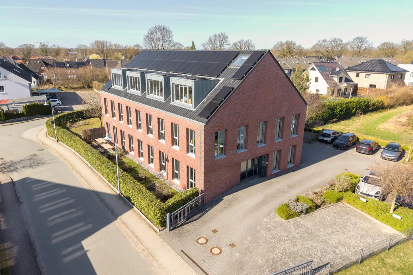 Aerial view of a modern red brick apartment building with solar panels on the roof, surrounded by a parking lot and trees.