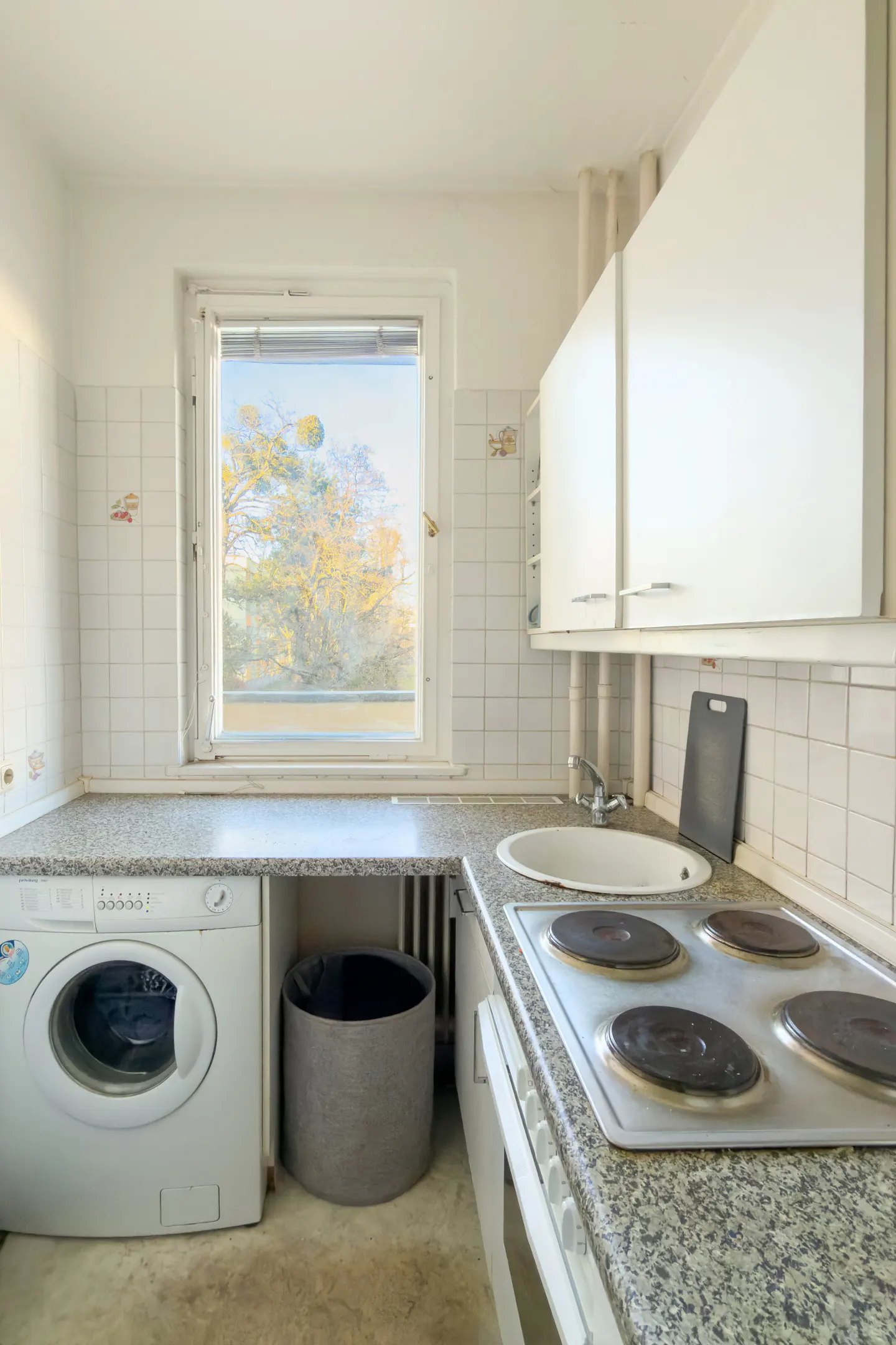 A bright kitchen with white cabinets, a washing machine, and a window showing trees.
