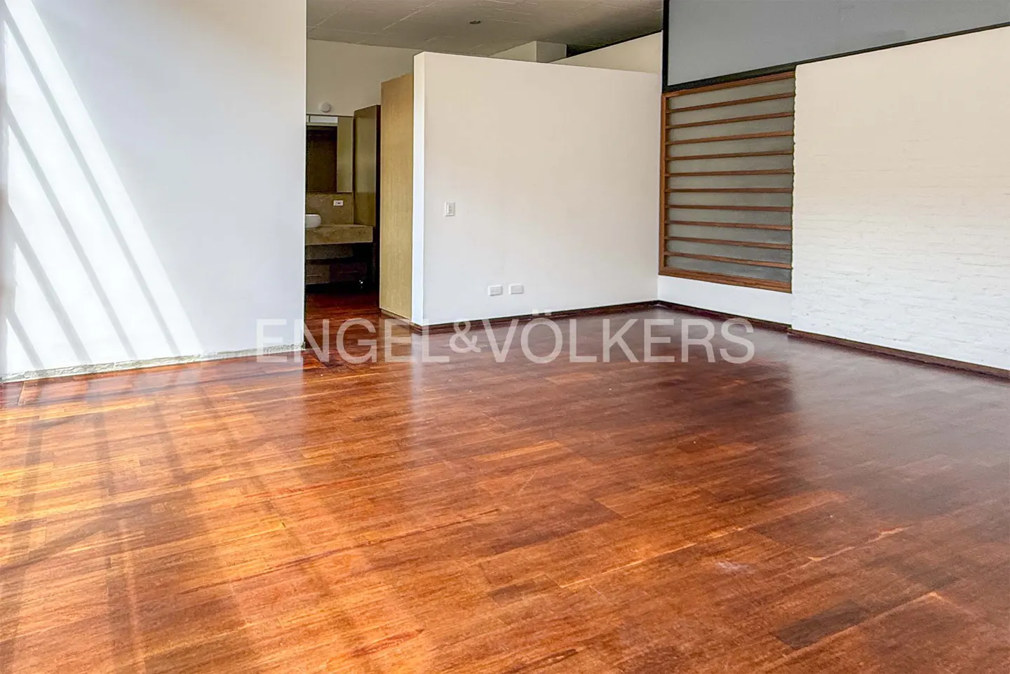 An empty room with a shiny, dark wood floor, white walls, and a glimpse of a bathroom in the background.