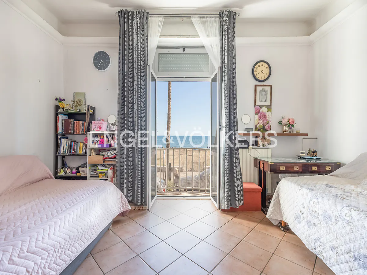 Bedroom with two beds, tiled floor, and open balcony doors revealing a beach and ocean view. Gray patterned curtains frame the doorway.