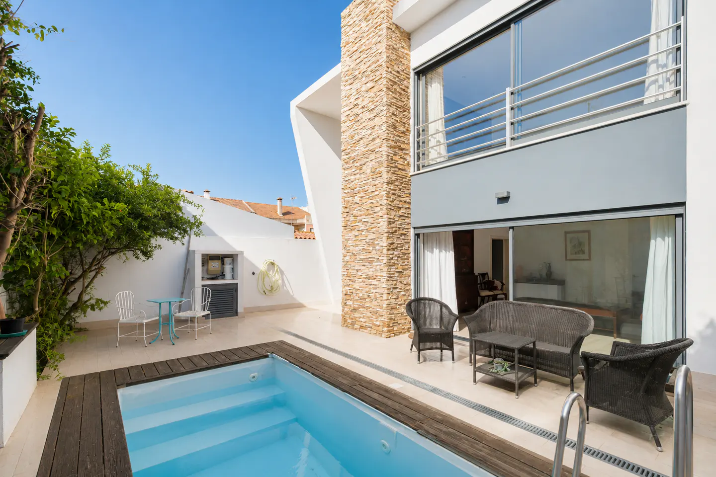 Modern home exterior with a small pool, stone accent wall, and a patio featuring wicker furniture under a clear blue sky.