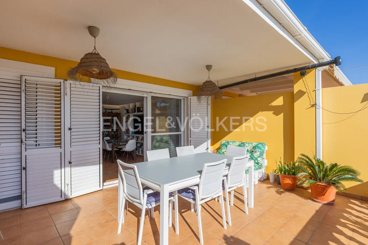 Covered patio with a white table and chairs, straw pendant lights, and yellow walls. Plants in pots line the back wall. Engel & Völkers logo on the sliding glass door.