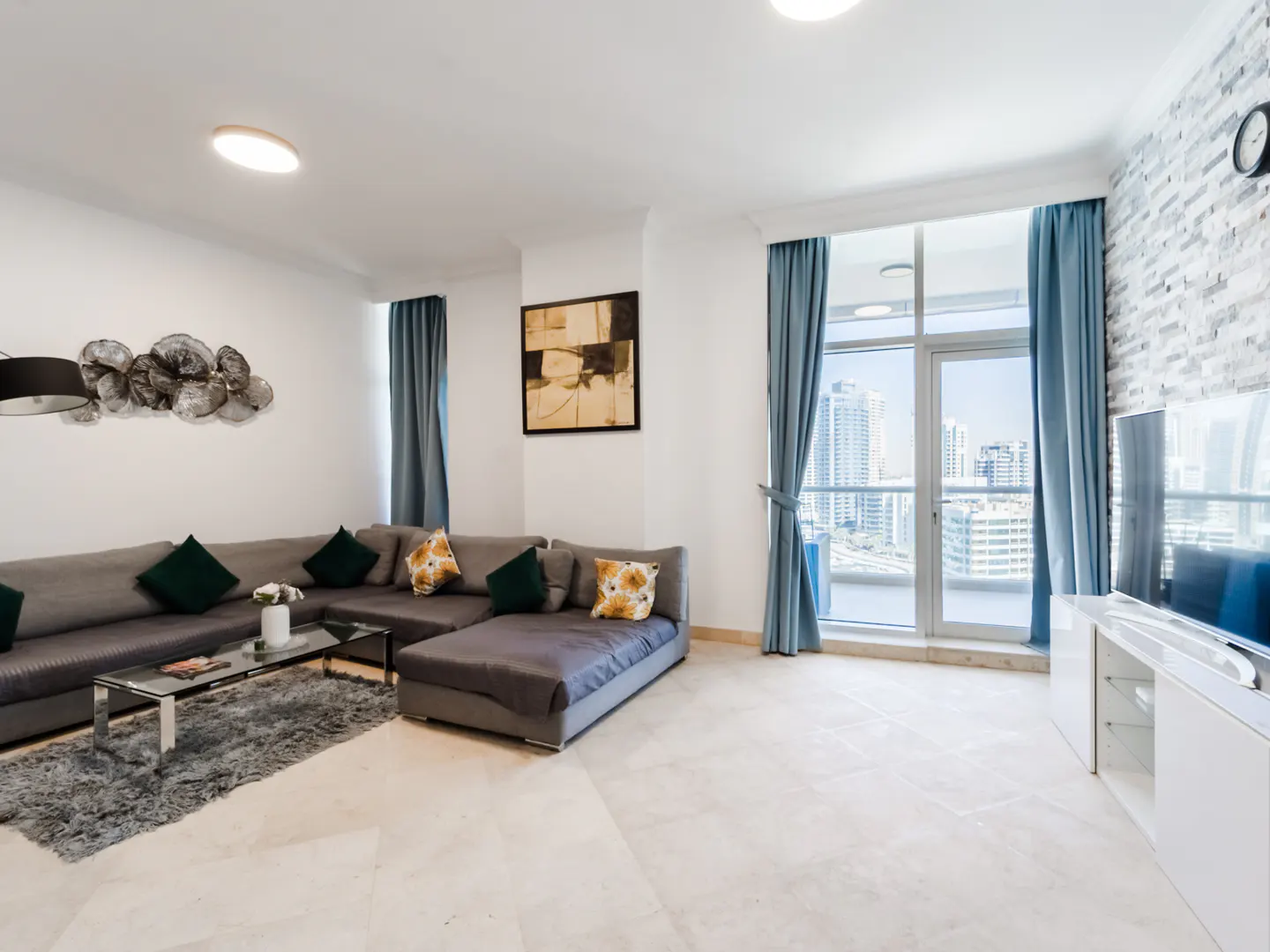 Modern living room with a grey sectional sofa, glass coffee table, and floor-to-ceiling windows overlooking a city skyline.