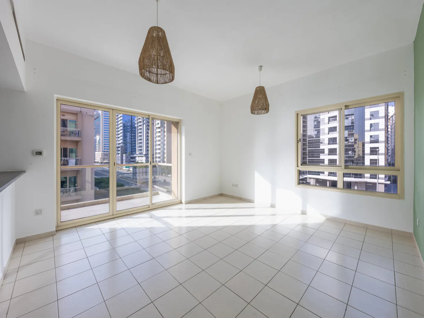 Bright, empty room with white walls, tile floor, and two woven pendant lights. Large sliding glass doors and a window offer city views.