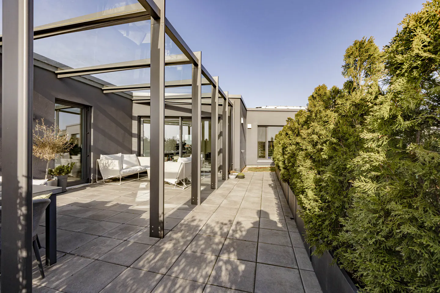 Modern rooftop patio with gray pavers, a glass-covered pergola, white seating, and a green hedge against a blue sky.