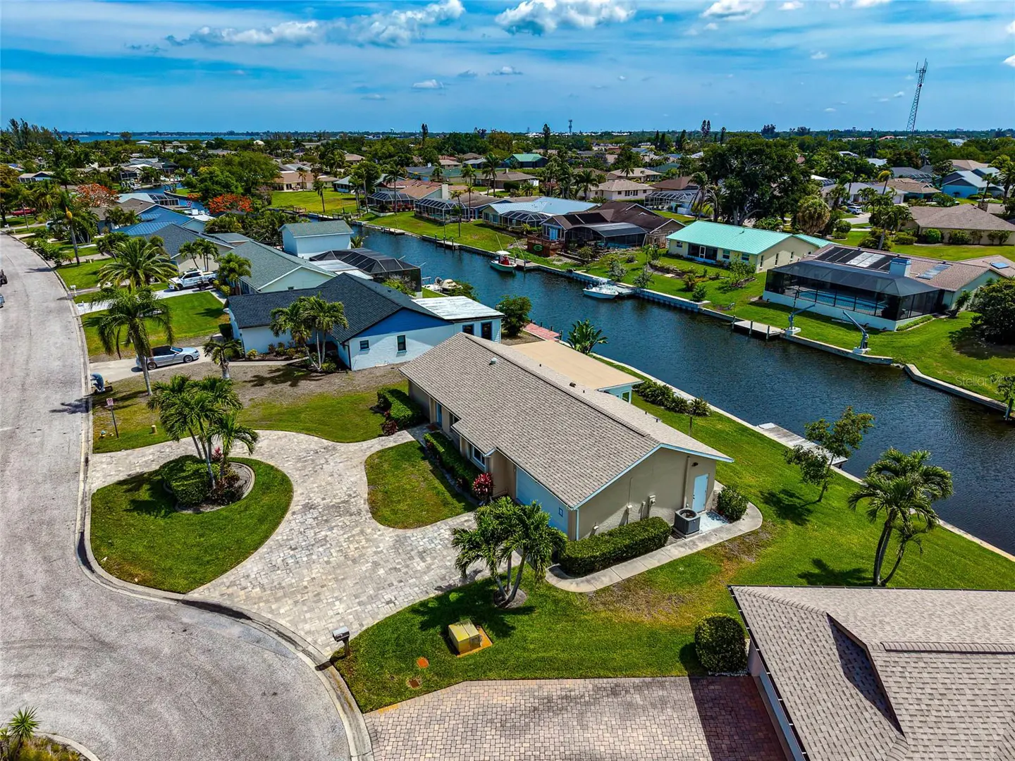 Aerial view of a beige single-story home with a gray roof, palm trees, and a canal in a residential neighborhood.