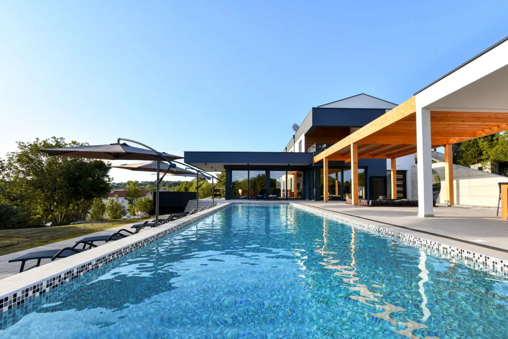 Modern home exterior with a long, blue tiled pool, lounge chairs, and umbrellas under a clear blue sky.