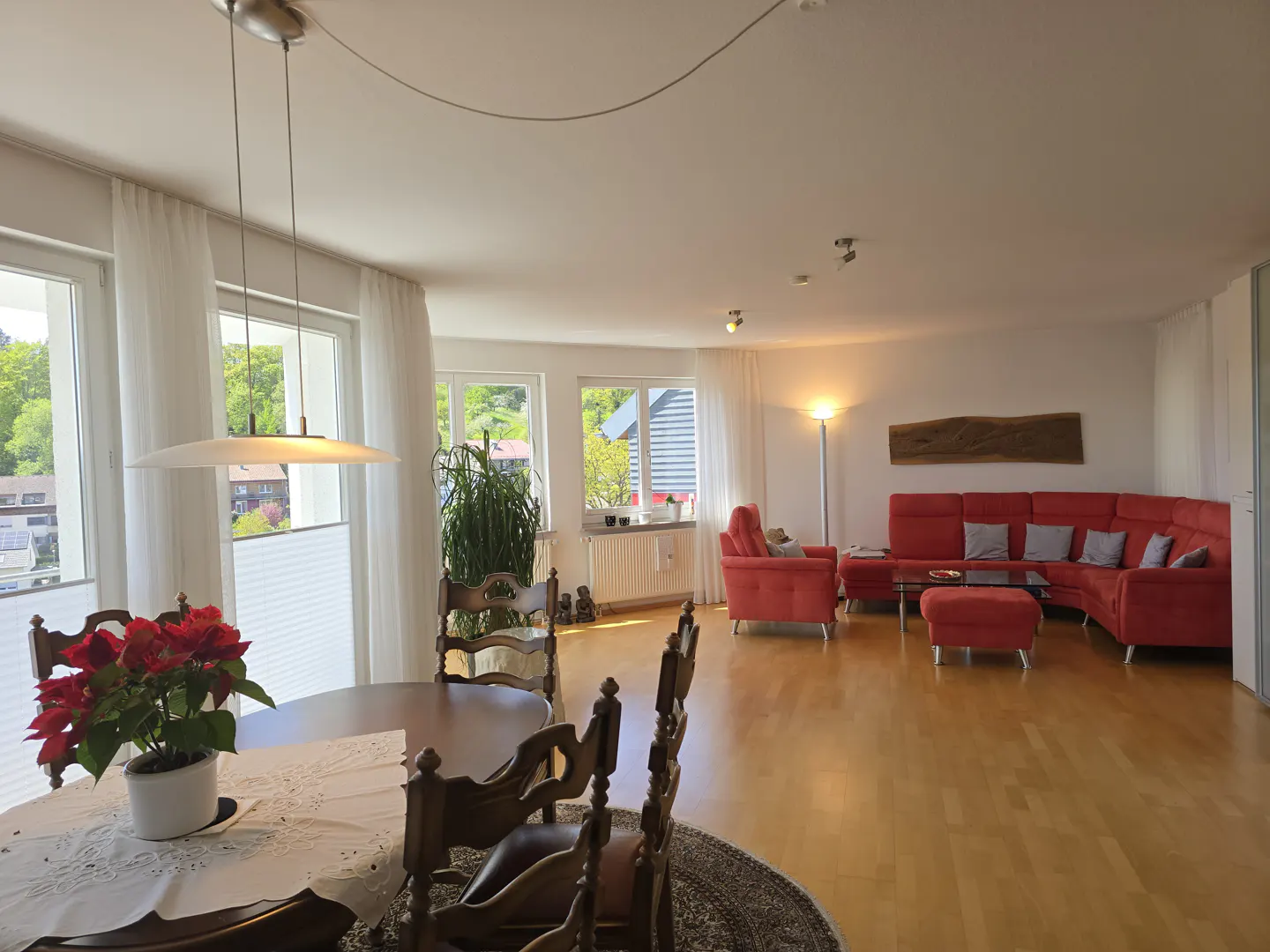 Bright, open-plan living and dining area with wood floors. A red sectional sofa and dining table with poinsettia are visible.