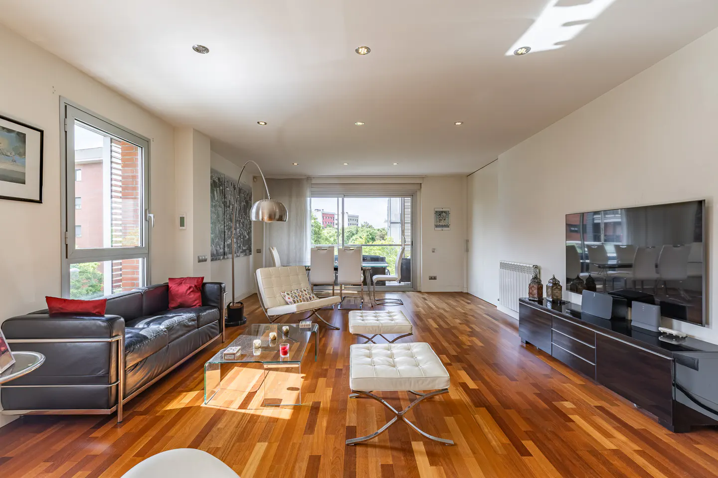 Bright living room with hardwood floors, black leather sofa with red pillows, white chairs, and a glass coffee table.