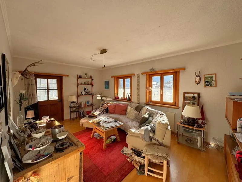 Cozy living room with a beige sofa, red rug, and wooden accents. Natural light streams through windows. Antlers hang above a door.